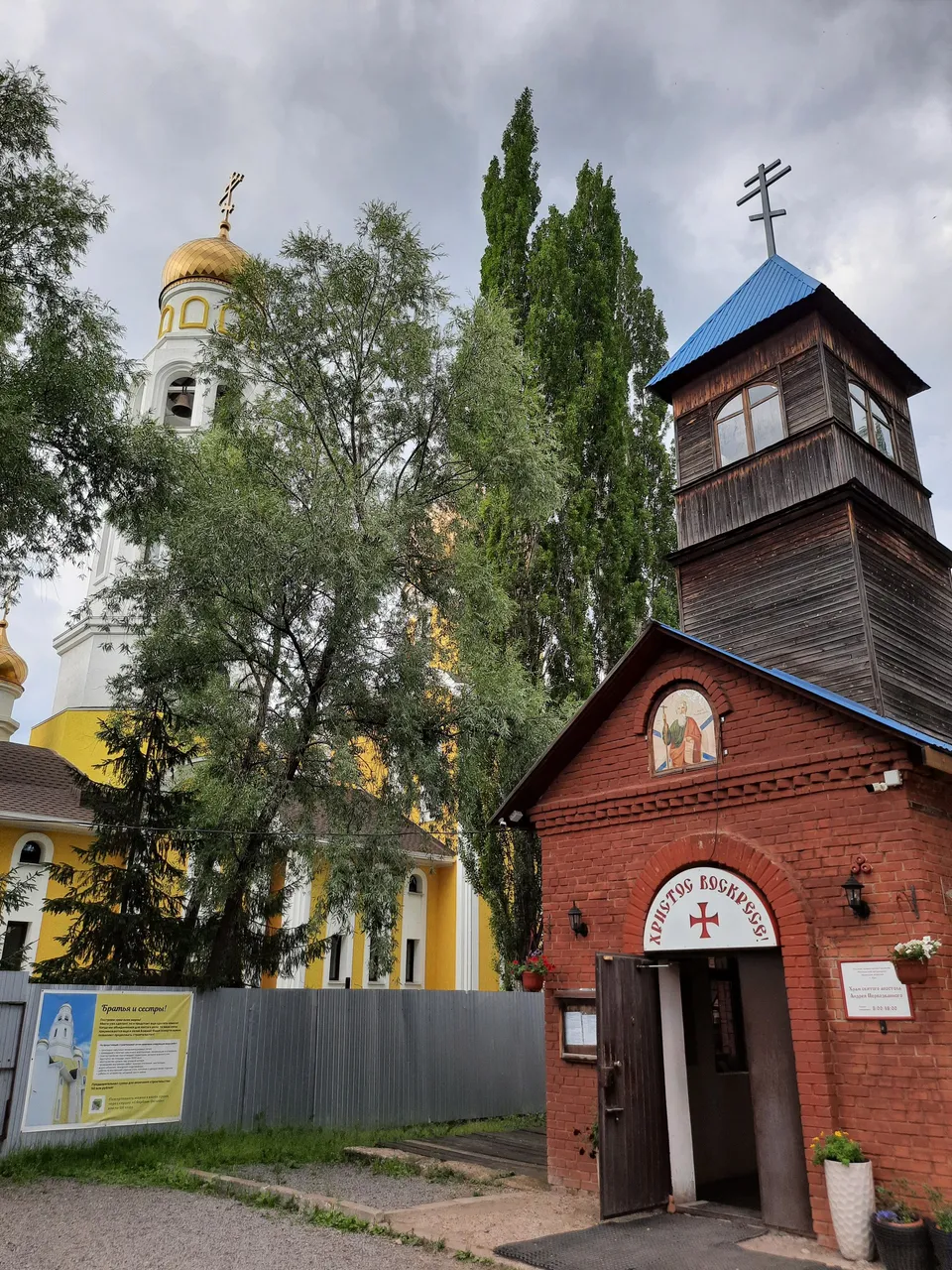 Старое кирпичное здание с тёмной деревянной звонницей, вывеска и крест над входом, рядом новый храм