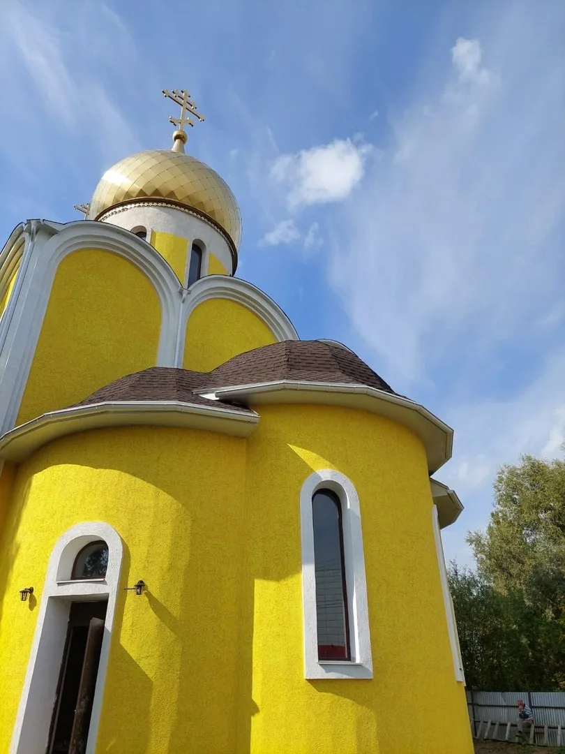 Золочёный купол с крестом и завершённая тёмная кровля на жёлто-белом строящемся храме на фоне облачного неба