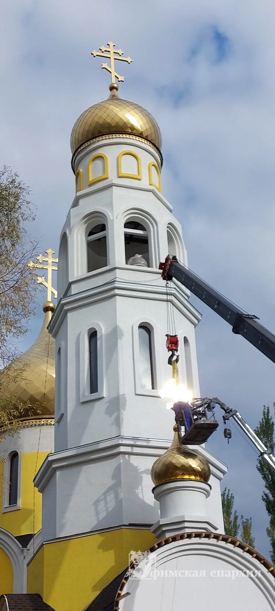 Звонница нового храма с золочёным куполом, кран рядом устанавливает малую главу